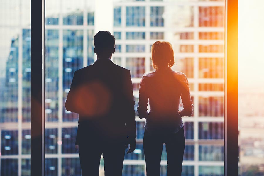 About Our Agency - Two Business Associates Stand in Silhouette Against a Large Window in a Skyscraper, Orange Sunlight Streaming in at Dusk