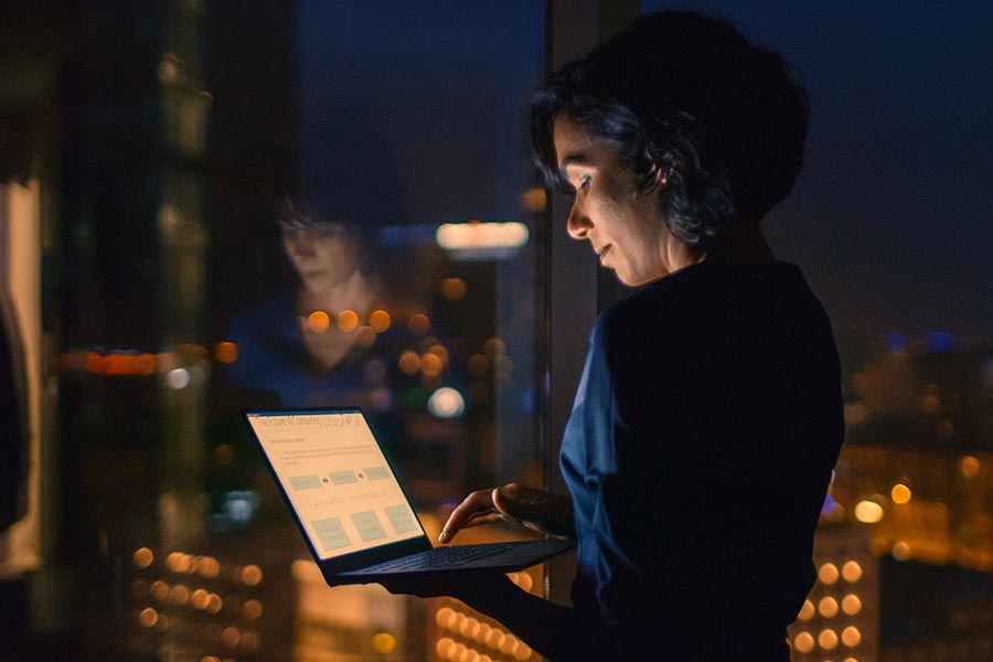 Client Center - Woman Uses a Computer Next to a Large Window at Night With City Lights Glowing Below
